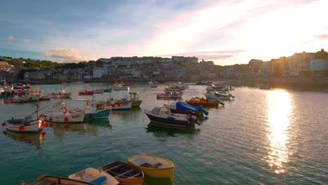Famous-seaside-coastal-tourist-destination-town-St-Ives-with-beach-and-harbor-promenade-in-Cornwall-in-Southern-England,-UK-with-fishing-boats-moving-in-the-clear-blue-water-by-sunset-and-reflections