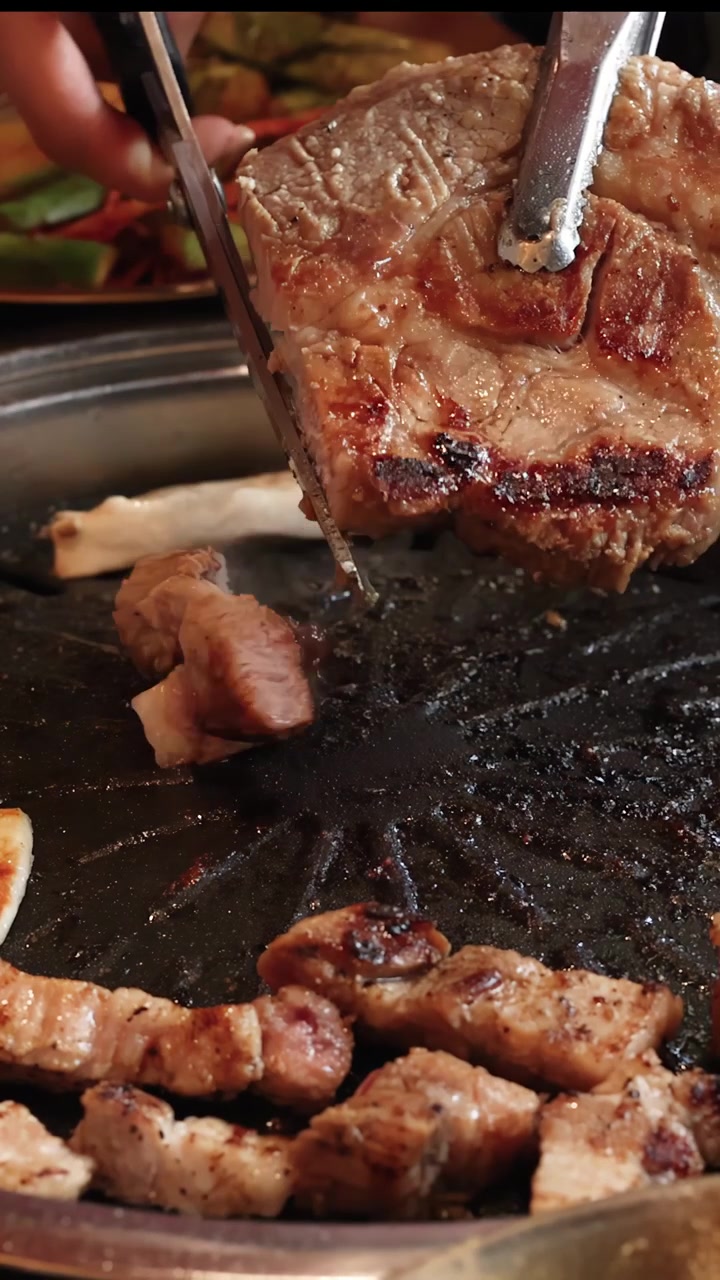Tangan menggunakan penjepit logam untuk memotong daging babi panggang di atas pemanggang barbekyu Korea meja, pencahayaan hangat.
