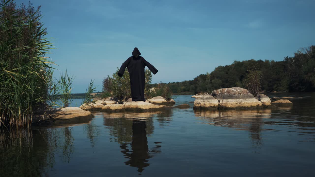 Evil witch in black cloak with hood on stones near the river. Divination by the water. Halloween costume.