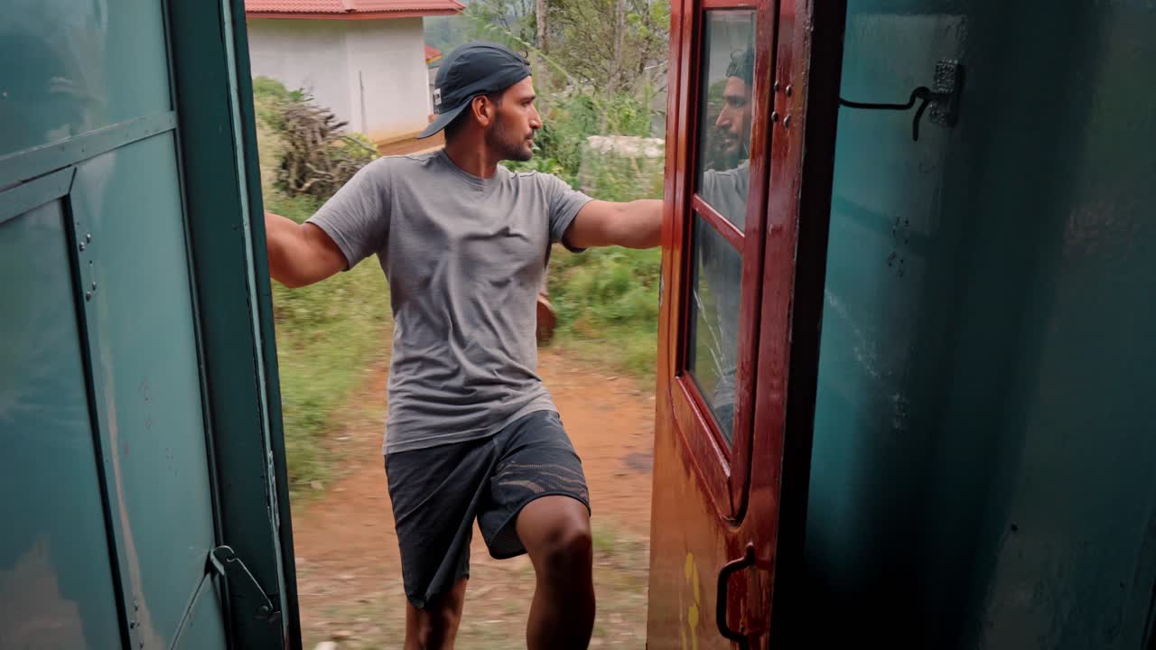 A man sits at the open door of a moving train on the scenic Ella to Kandy route in Sri Lanka, peacefully enjoying the lush landscape of tea plantations and rolling hills.
