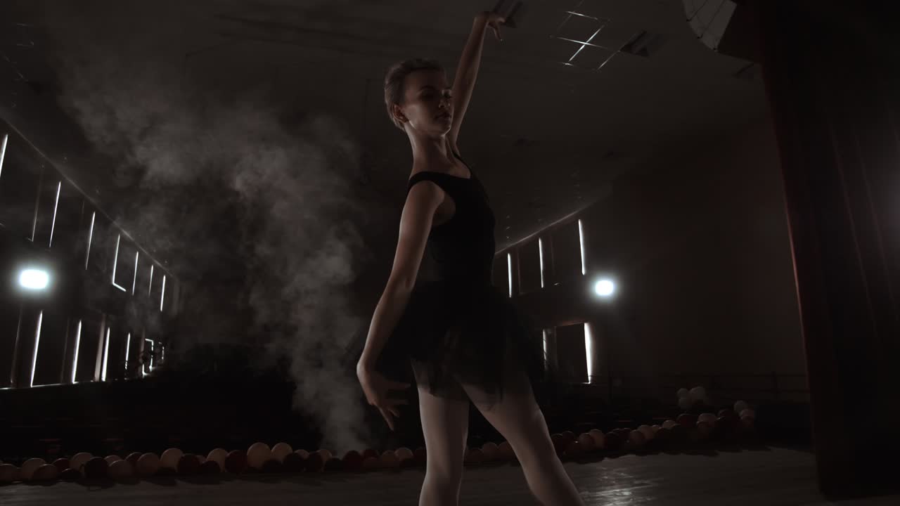 A graceful ballerina dancing ballet elements in the dark with light and smoke on the background, slow motion.