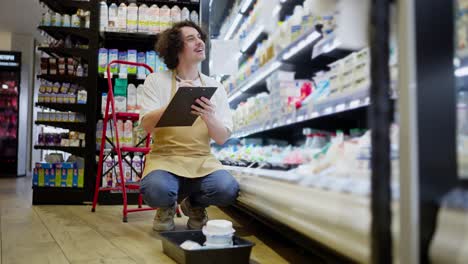 Ein-Glücklicher-Brünetter-Typ-Mit-Schnurrbart-In-Einer-Schürze-Verwendet-Ein-Tablet,-Um-Waren-In-Der-Milchabteilung-Eines-Supermarkts-Nachzuzählen