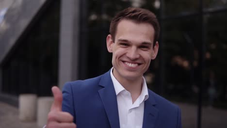 Upwards-footage-of-a-young-businessman-in-elegant-blue-suit-standing-outdoors,-cheerfully-smiling-and-showing-thumb-up.-Handsome