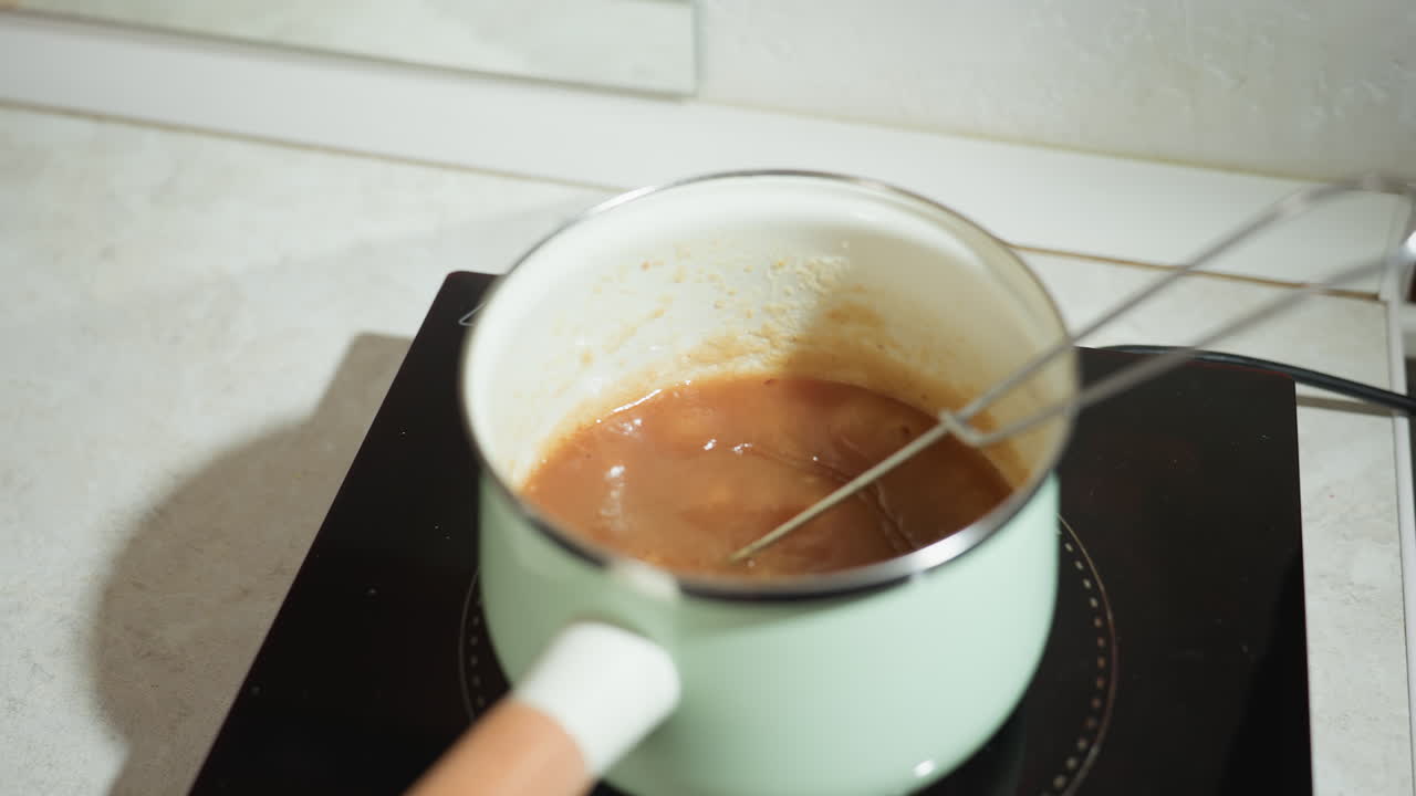 Soup steaming on electric stove in light green pot with metal whisk resting inside, thick brown mixture bubbling from heat, countertop
