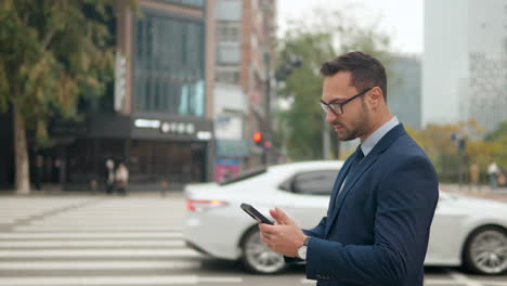 Geschäftsmann-Hält-Telefon-In-Der-Hand,-Während-Er-Auf-Dem-Bürgersteig-Steht,-Mit-Verkehr-Im-Hintergrund-In-Seoul,-Südkorea