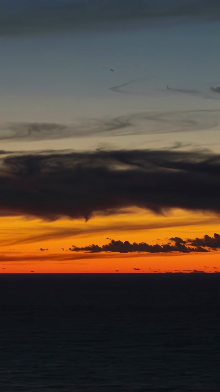 Shifting dark cloud band and orange horizon deepening over coastal sea, showing tendril and specks