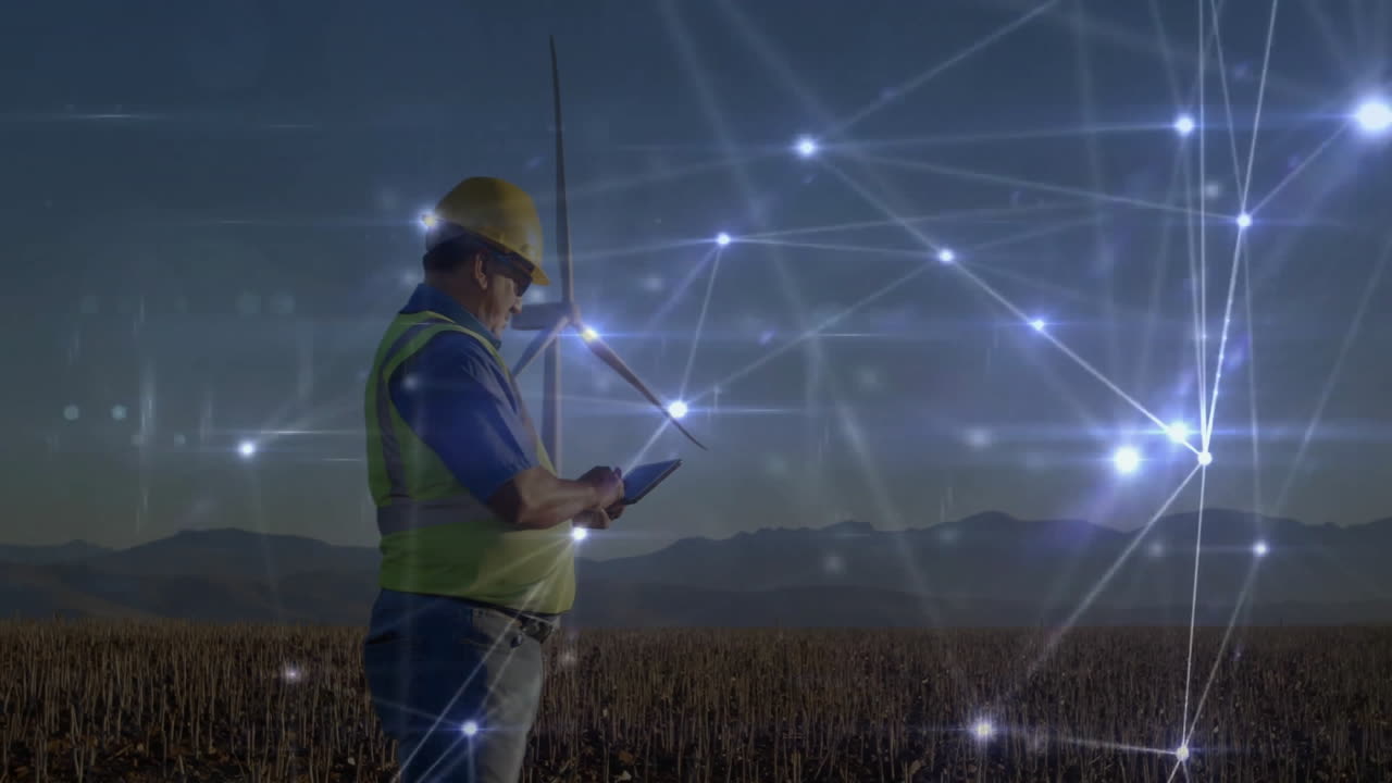 Male technician using tablet in wind farm at dusk, showcasing energy technology network graphics
