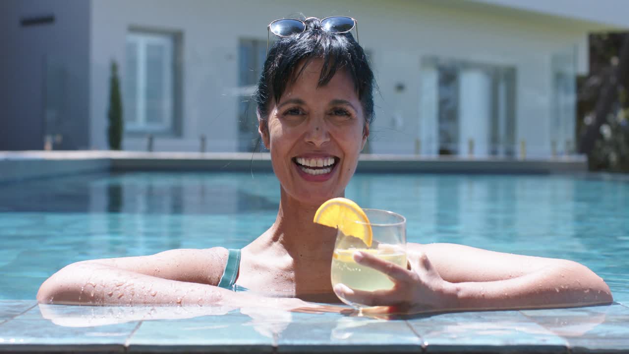 Camera rolling, smiling woman in green swimsuit and shades, holding citrus drink, laughing at pool