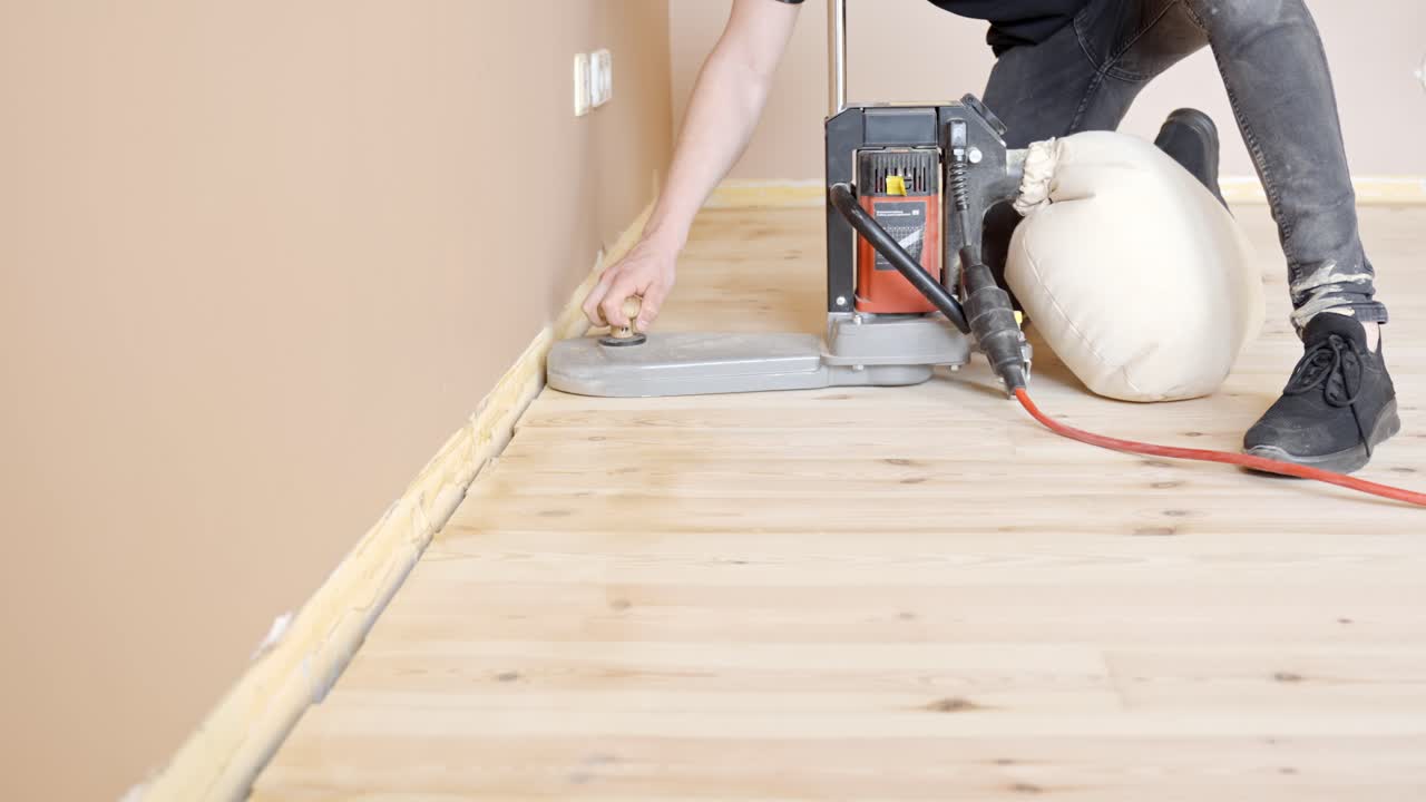 Person sanding wooden floor in empty bare room, smoothening the surface with specialized tools, flooring is light colored, modern and new