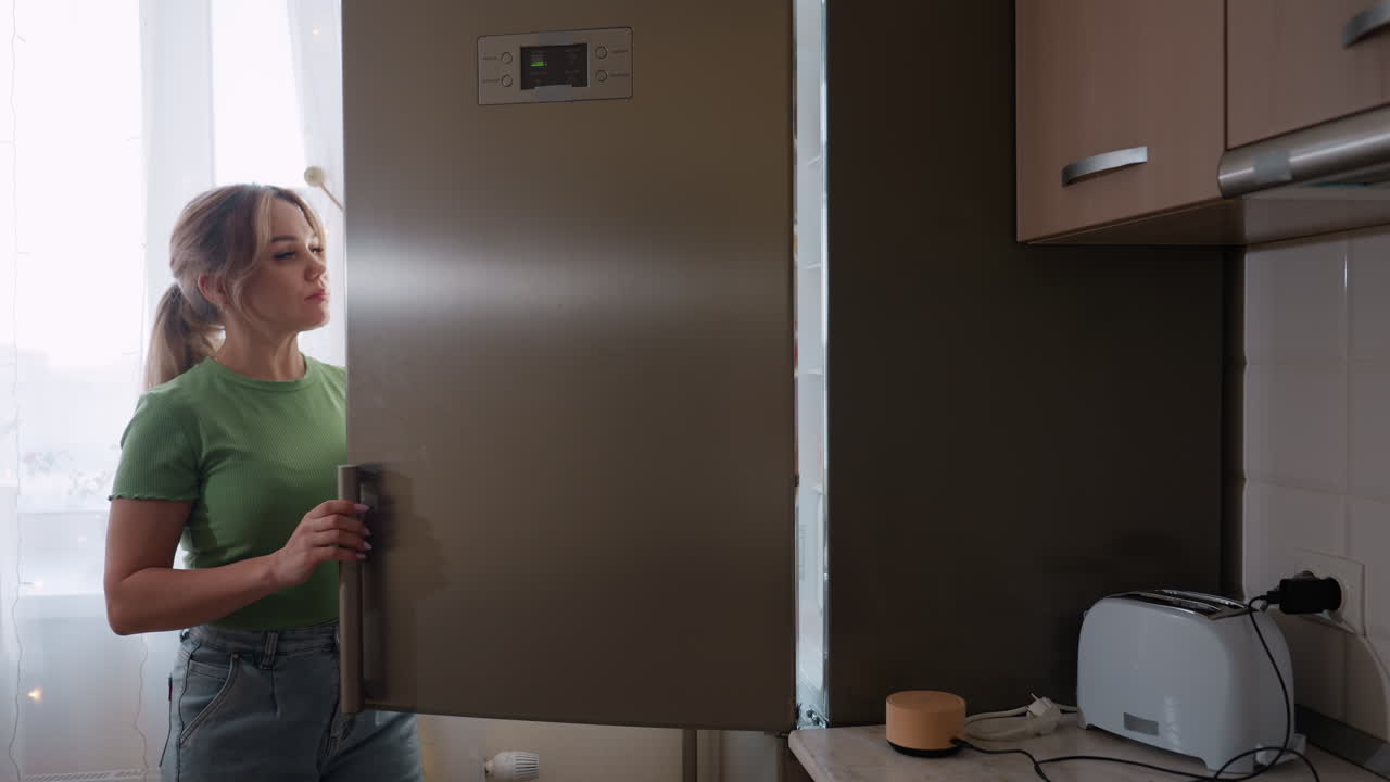 Side view of woman in green shirt retrieving eggs from refrigerator in kitchen, preparing ingredients for cooking while standing near counter and cabinets in bright domestic interior