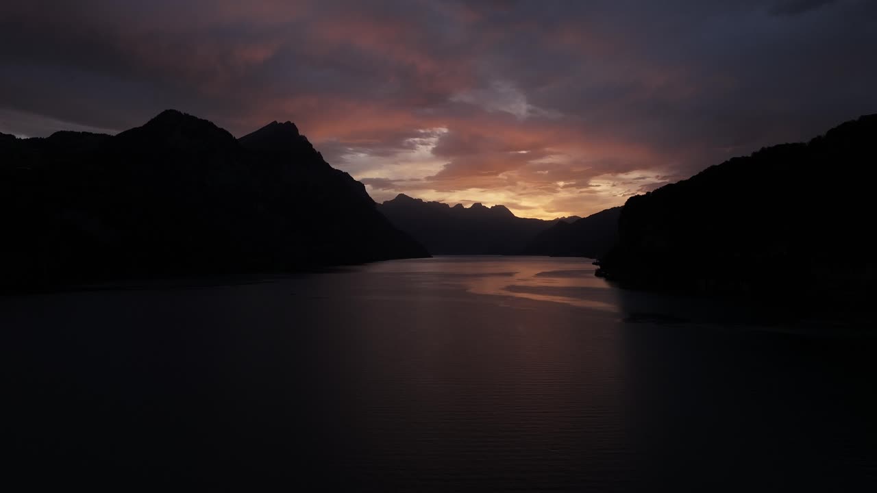 Drone slowly pushes toward Walensee lake, warm orange light of morning sunrise illuminating clouds above dark mountains, stunning landscape view in Wessen, Quinten, Switzerland.