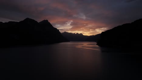 drone slowly pushes toward walensee lake, warm orange light of morning sunrise illuminating clouds above dark mountains, stunning landscape view in wessen, quinten, switzerland.