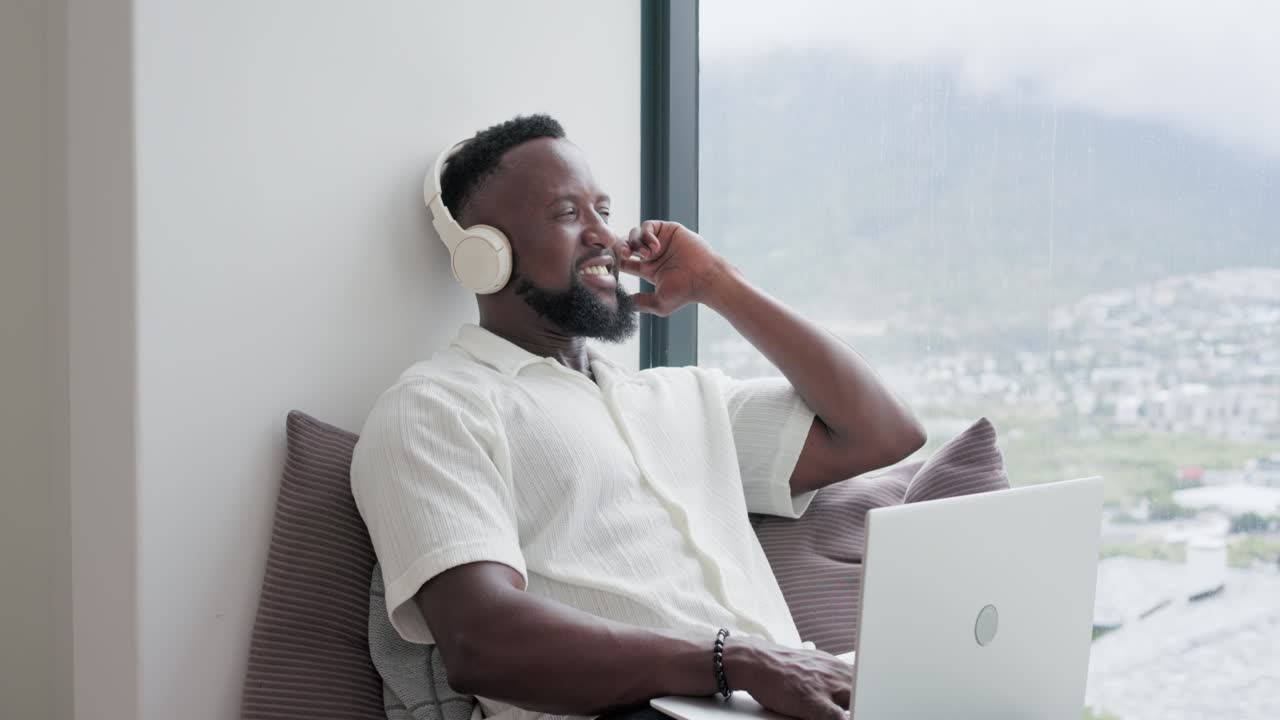 After pausing typing, man is adjusting headphones and singing along to music on laptop by window