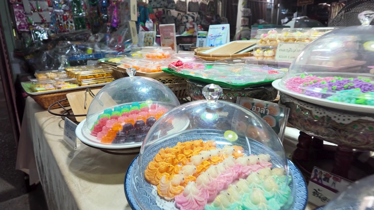 Close-up of vibrant flower-shaped Thai sweets under glass domes at a Koh Kret market stall.