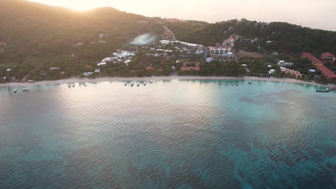 Veduta aerea cinematografica della costa caraibica con barche, acqua turchese, spiaggia di sabbia bianca e resort, all'alba, con l'isola di Roatan, West End, Honduras, sullo sfondo