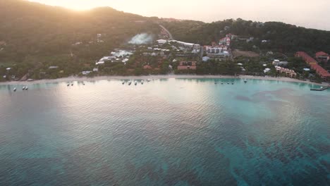 vista aérea cinematográfica de la costa caribeña con botes, agua turquesa, playa de arena blanca, resorts durante el amanecer, fondo de la isla, isla de roatán, extremo oeste, honduras