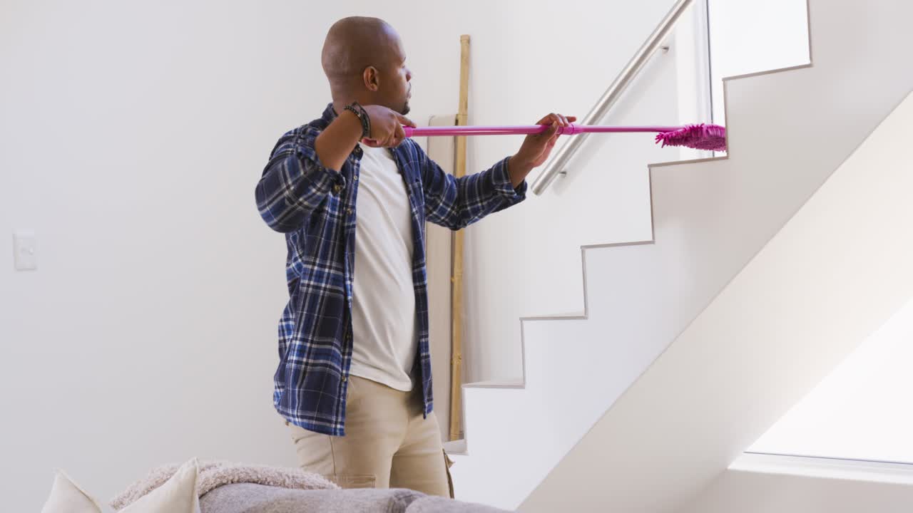 African American man spotting dust, using long pink mop, reaching, removing dust on home handrail