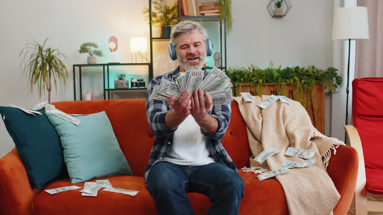 무선 헤드폰을 착용한 행복한 백인 노인이 돈을 비하는 것을 보여줍니다.