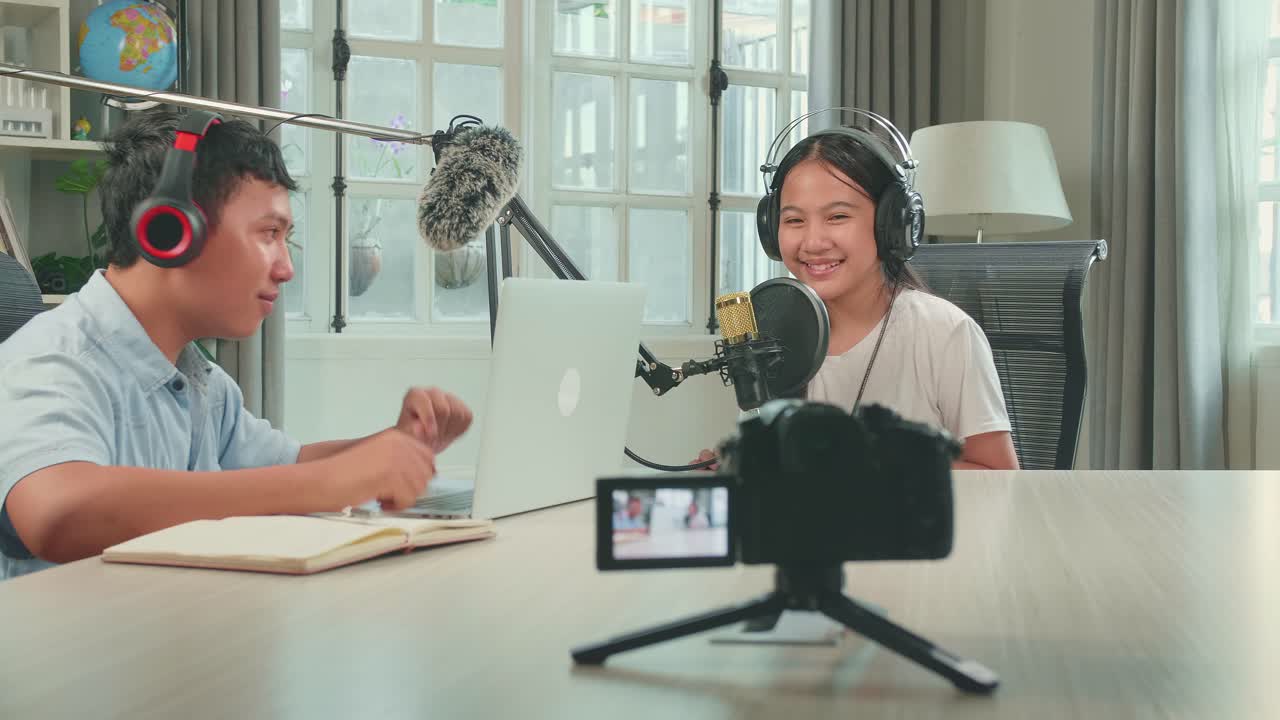 Camera Recording Of Asian Boy Host Record Podcast Use Microphone Wear Headphone With Laptop Interview Guest Conversation In His Home Studio, They'Re Talking And Looking At Camera For Content. Content Creator Concept