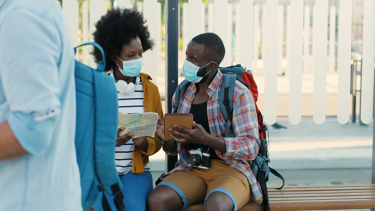 African american young happy man and woman travellers in facial masks sitting at bus stop talking and watching a map and a tablet to plan a route