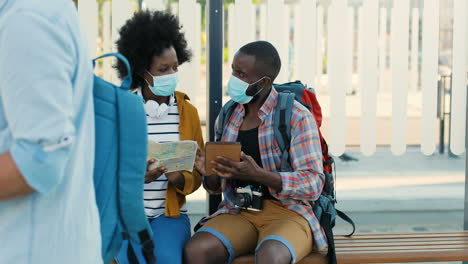 african american young happy man and woman travellers in facial masks sitting at bus stop talking and watching a map and a tablet to plan a route