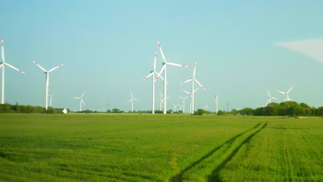 Parque-Eólico-Panorámico:-Vista-Desde-La-Ventana-Del-Tren-En-Cámara-Lenta,-Con-Numerosos-Aerogeneradores-Contra-El-Cielo-Azul-Y-Campos-Verdes