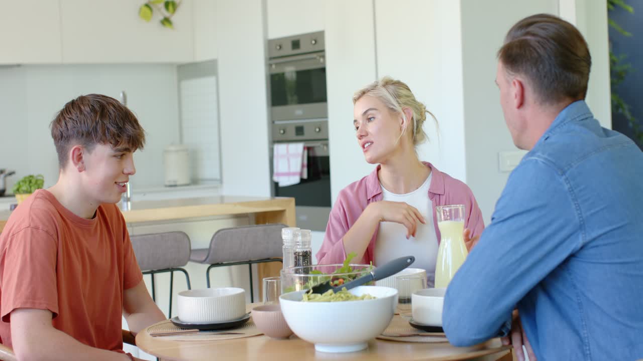 Familia alrededor de la mesa redonda de la cocina, el padre sosteniendo un gran tazón blanco de ensalada y sirviendo, la madre animando al adolescente a hablar.