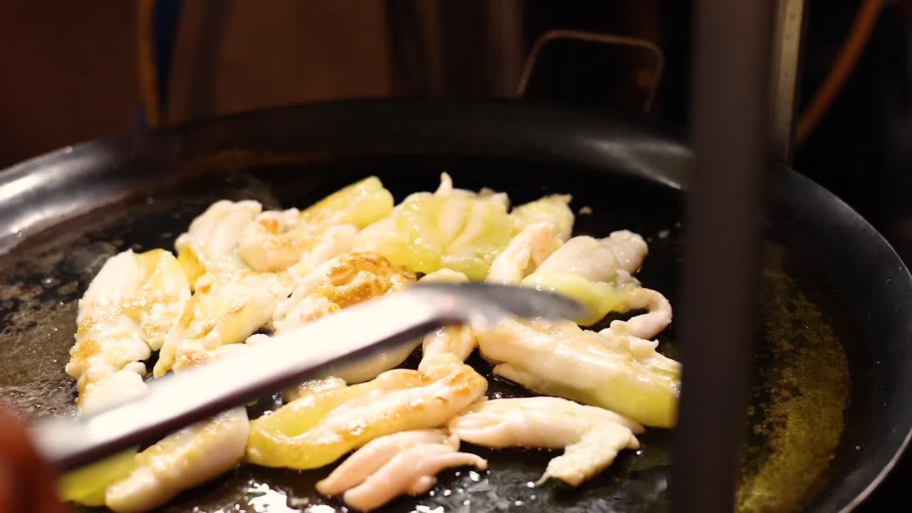 Hands use tongs to fry squid eggs on large griddle under warm street food lighting