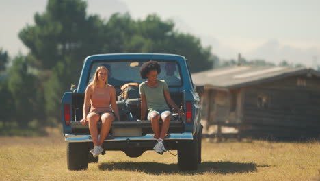 Zwei-Frauen-Fahren-Auf-Dem-Rücksitz-Eines-Pick-up-Trucks,-Während-Freunde-Auf-Einem-Roadtrip-In-Einer-Hütte-Auf-Dem-Land-Ankommen