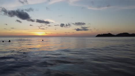 Luftaufnahme:-Drohne-Fliegt-Supertief-über-Den-Küstenwellen,-Menschen-Schwimmen-Im-Sonnenuntergang-Im-Meer-Mit-Inseln-Im-Hintergrund