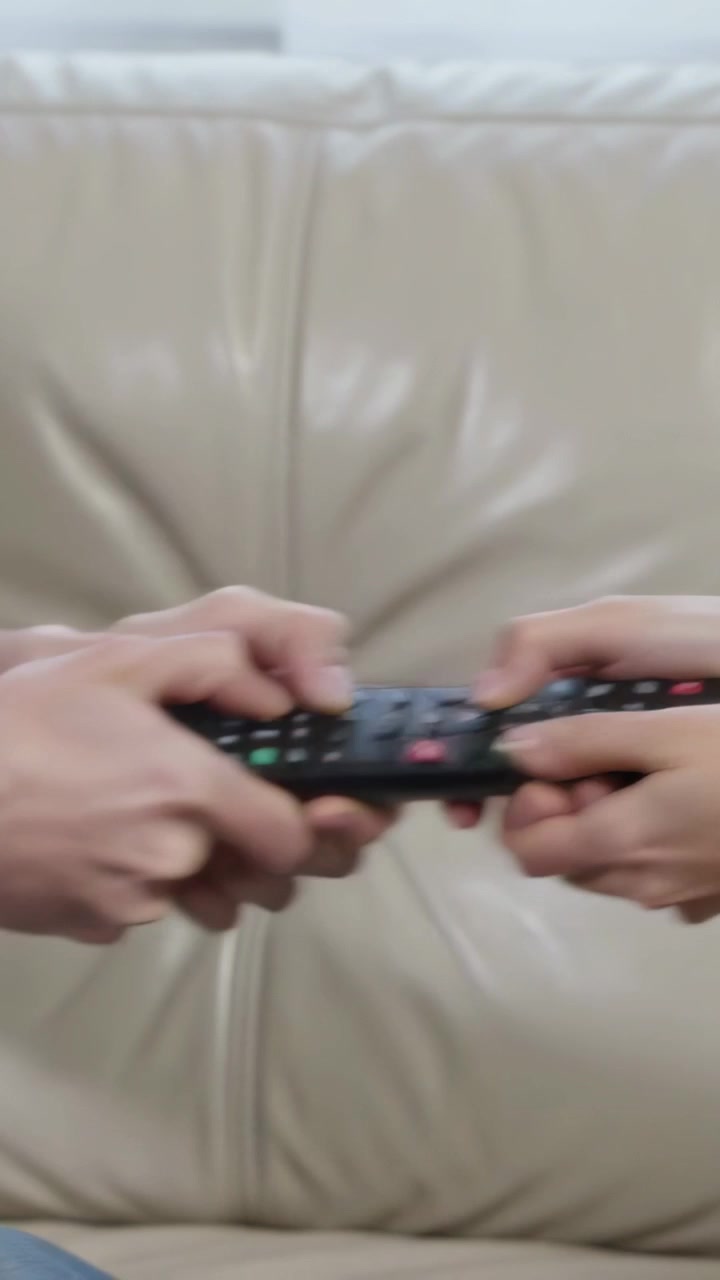 Video verticale: Due mani con maglia a maniche lunghe bianca che tirano un telecomando nero su un divano beige, in contesa
