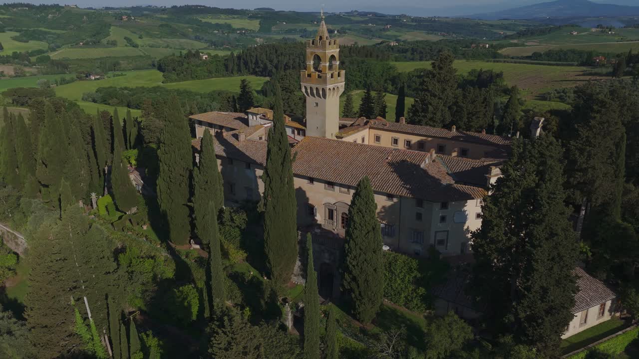 Drone shot flying directly towards Castello di Montegufoni in Tuscany. The historic castle and its iconic tower rise above cypress trees, surrounded by the lush green landscape of Florence’s hills.