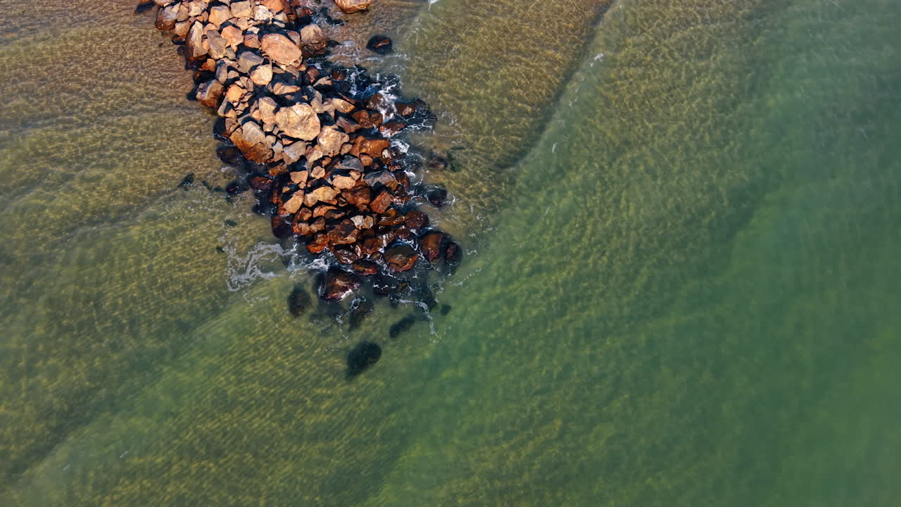 Rock formations extend into calm water at the beach. Gentle waves lap against the rocks. The sun shines brightly, creating clear visibility of the water