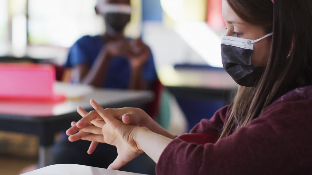 diversos escolares con máscaras faciales desinfectando las manos mientras están sentados en el aula.