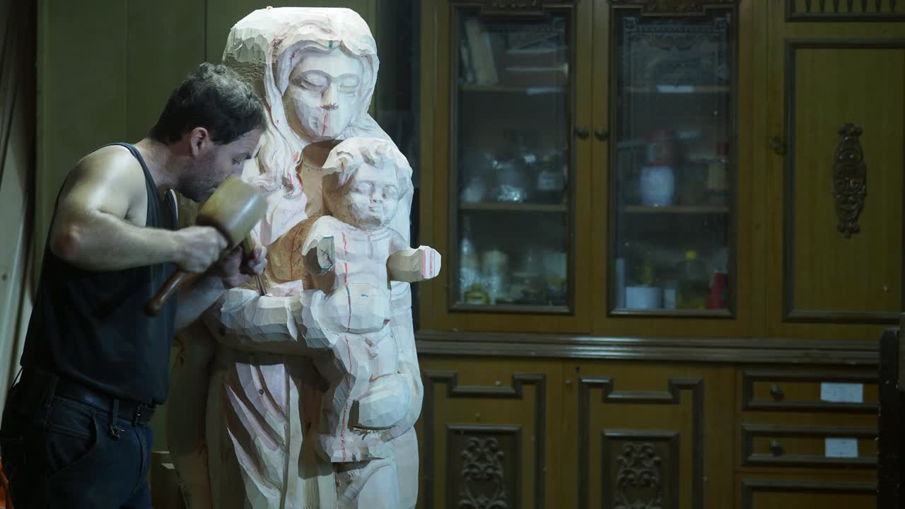 Caucasian adult craftsman chisels detail into rough wooden Madonna and Child sculpture, revealing veil folds and facial features against a workshop backdrop with storage cabinet, static camera