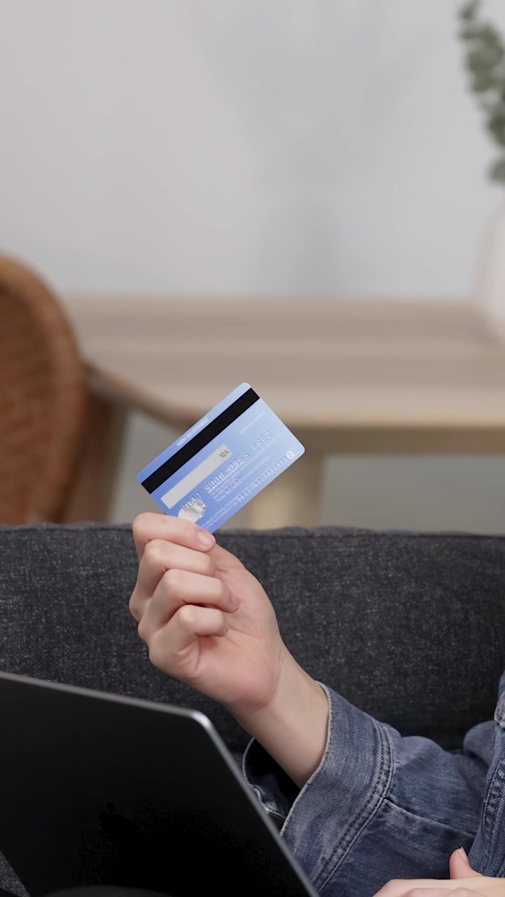 Donna asiatica sorridente seduta su un divano che usa un laptop e una carta di credito per il pagamento.