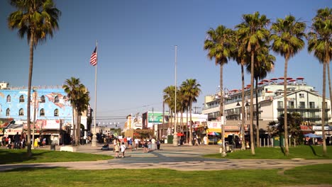 4k panning of venice beach in los angeles, park with green grass, palm trees, buildings and usa flag in the background, young people walking and scooters