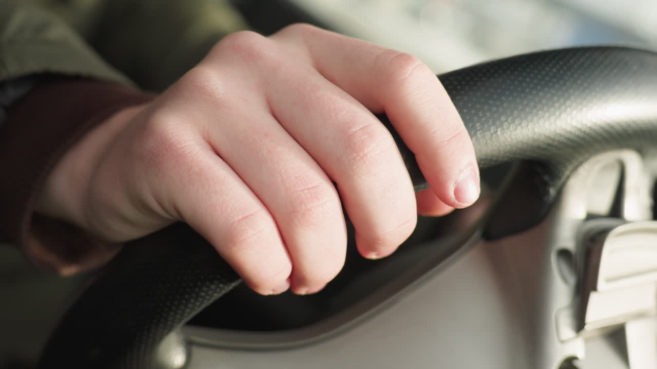 close up of hand resting on steering wheel gently tapping it while seated inside car with natural daylight filtering through window casting soft highlights on skin and interior details