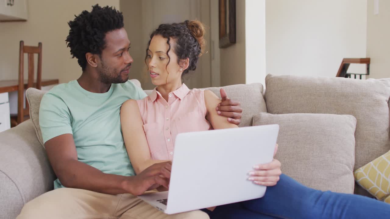 feliz pareja biracial sentada en el sofá con una computadora portátil y hablando
