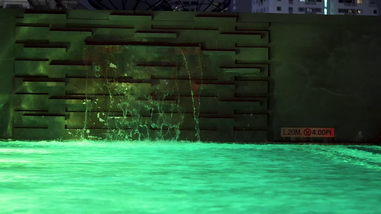 Illuminated rooftop pool with cascading waterfall, cityscape background, steady camera, nighttime ambient lighting