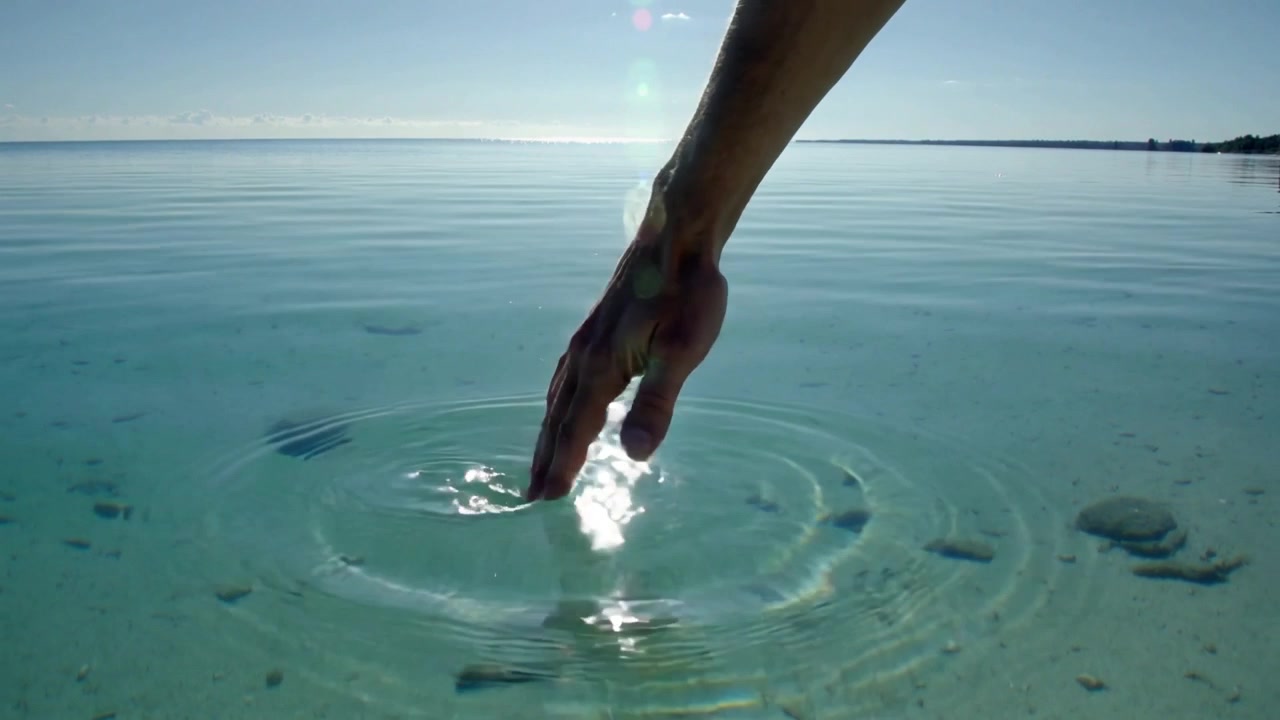 Reaching bare right hand touching and skimming shallow bay water, creating sun glare ripples