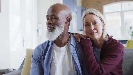 Portrait-of-happy-senior-diverse-couple-in-living-room-sitting-on-sofa,-embracing-and-smiling