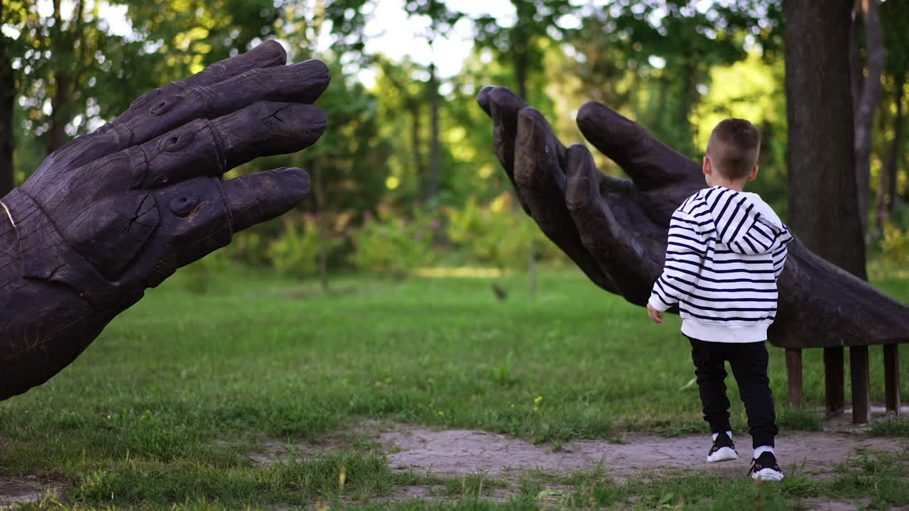 Three-year-old toddler standing at the sculpture of big wooden hands in the park. Kid checks the both sculptures and leaves.