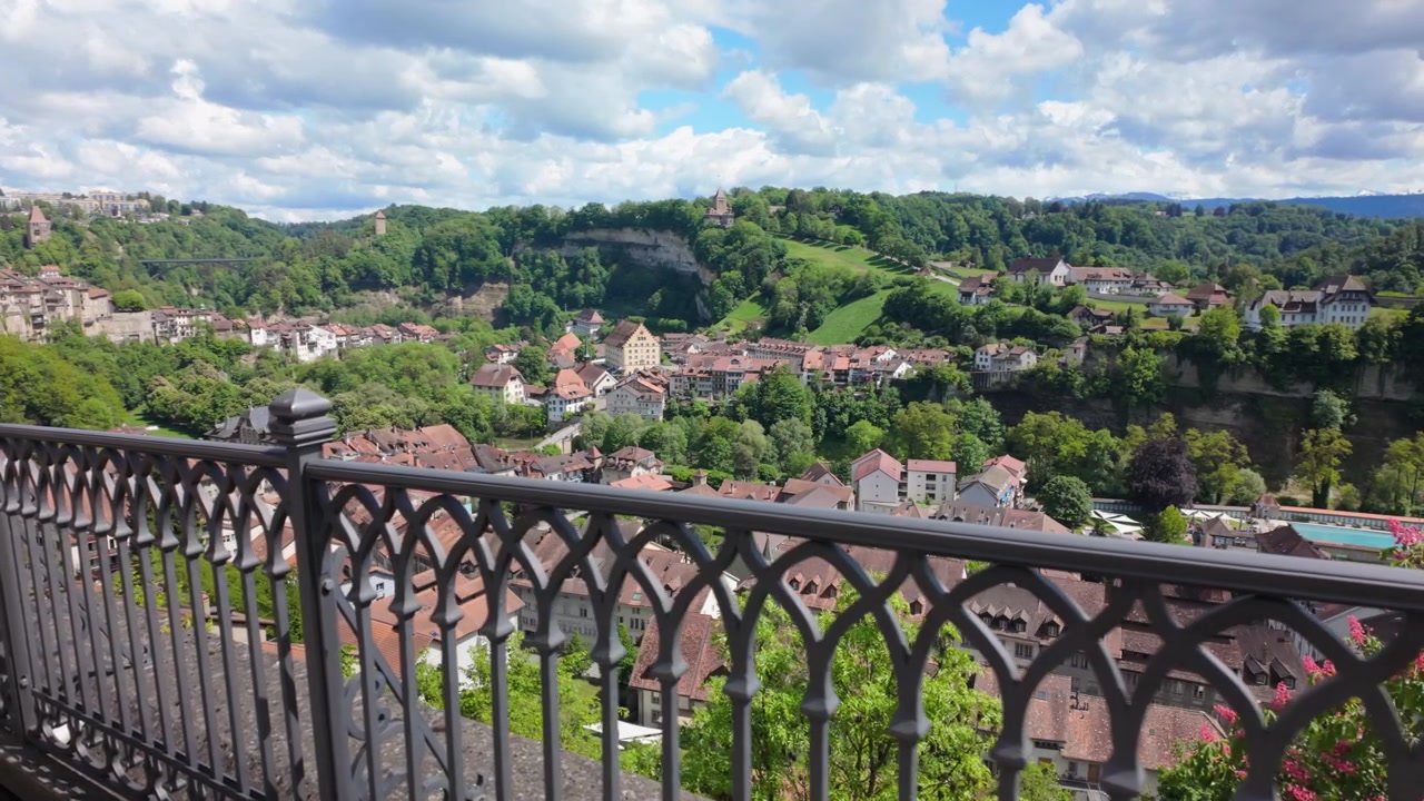 Berjalan di sepanjang pagar logam dengan pemandangan panoramik kota bersejarah Fribourg di Swiss, yang terletak di lembah hijau, dengan atap genteng merah, perbukitan, dan langit biru berawan, terlihat dari titik pandang yang tinggi.