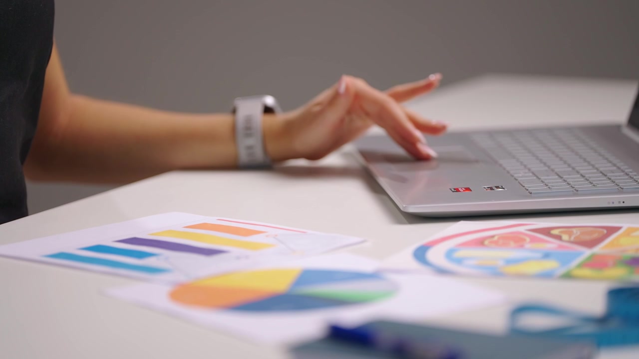 Primo piano di mani femminili che sistemano grafici e diagrammi nutrizionali colorati su una scrivania mentre usano un mouse del computer accanto a un laptop, mostrando la pianificazione dietetica e il lavoro professionale nel campo del benessere.