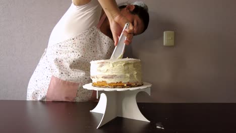 Latin-woman-wearing-an-apron-preparing-cooking-baking-a-cake-spreading-butter-frosting-with-a-white-plastic-cake-scraper