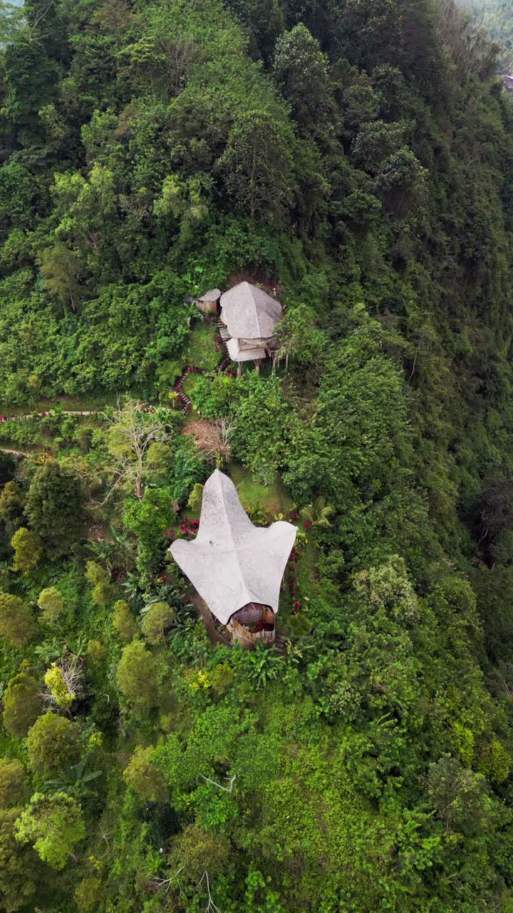 Vertical overhead view capturing a bamboo house on a tropical hillside in Sidemen, Bali. The overhead shot reveals the villa’s curved bamboo roof and its harmony with the jungle landscape