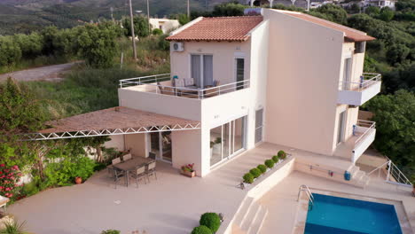 aerial pan shot of luxury greek villa with pool, sun loungers - outdoor seating nestled amongst green foliage