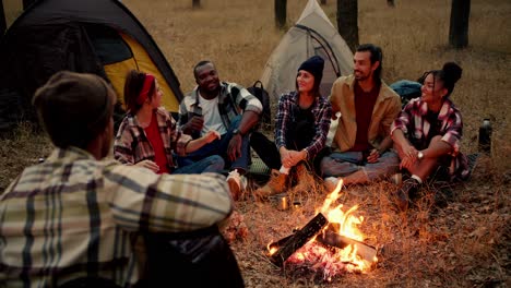 A-group-of-participants-in-a-group-hike-sit-around-a-fire-with-red-coals-and-communicate.-Girls-and-boys-sit-in-a-circle-around-the-fire-enjoying-the-view-and-singing-a-song-to-a-guitar-played-by-a-man-in-a-green-checkered-shirt