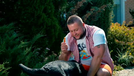 a young man feed a black dog a carrot in the courtyard